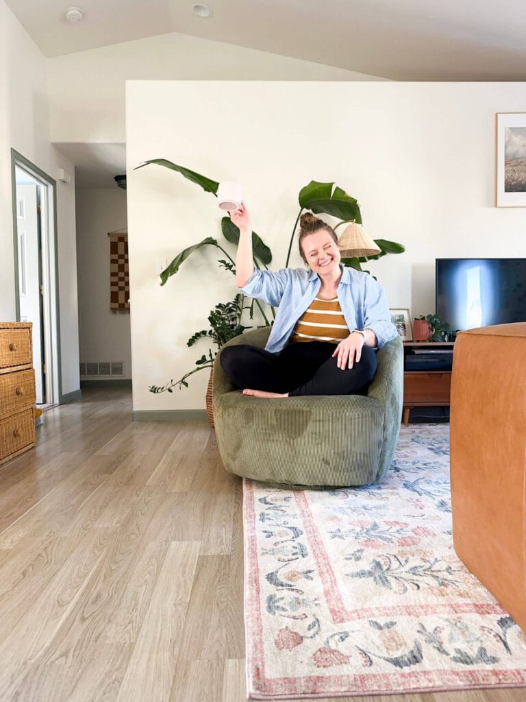 smiling woman sitting in green accent chair holding pink mug with arm raised