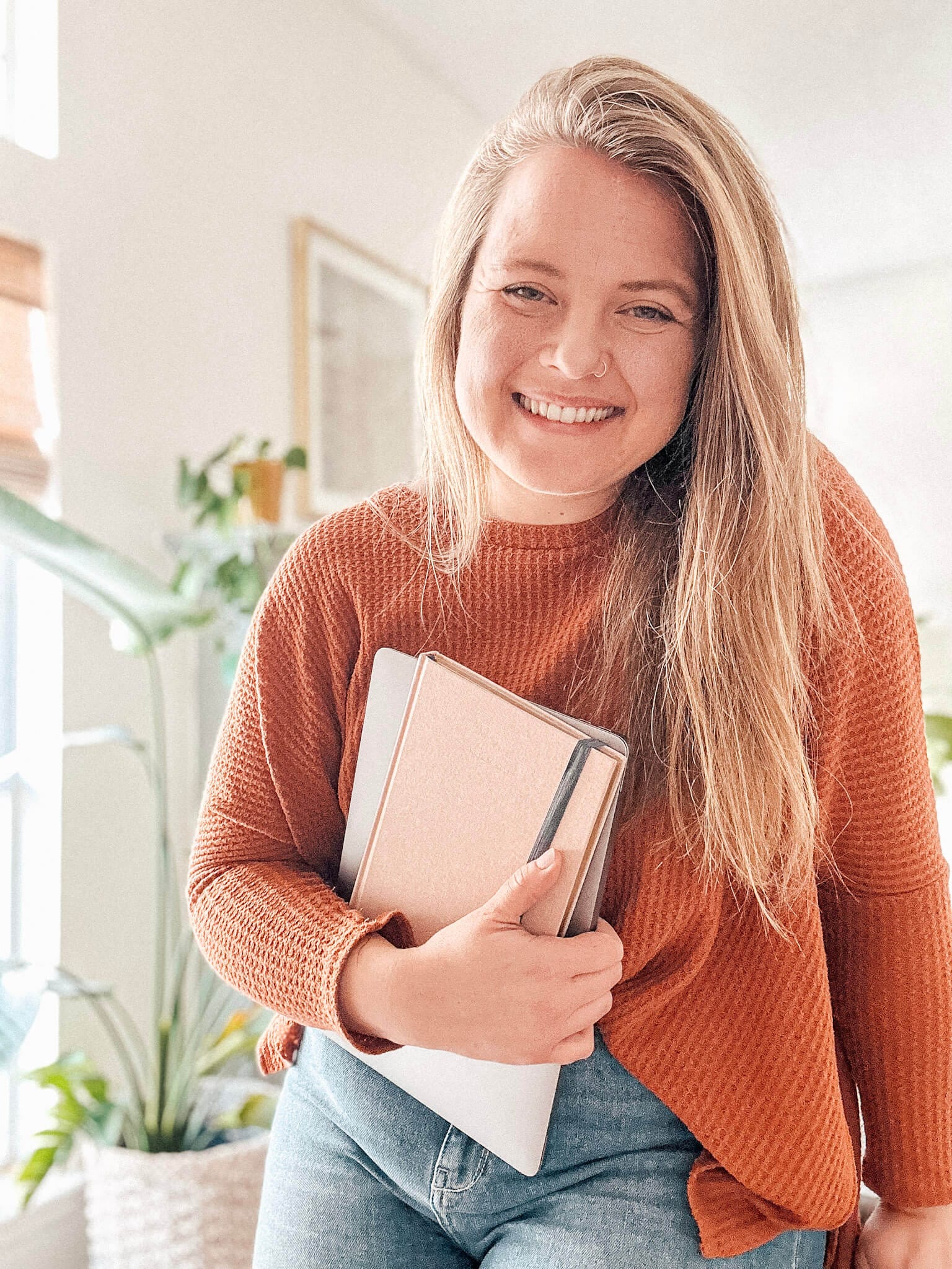 Woman standing in bright living room in orange long-sleeve holding a pink notebook