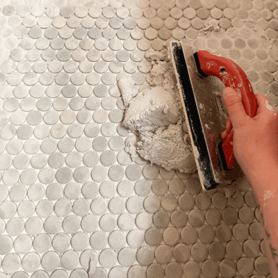 hand holding grouting tool with light gray grout being applied over green penny tile