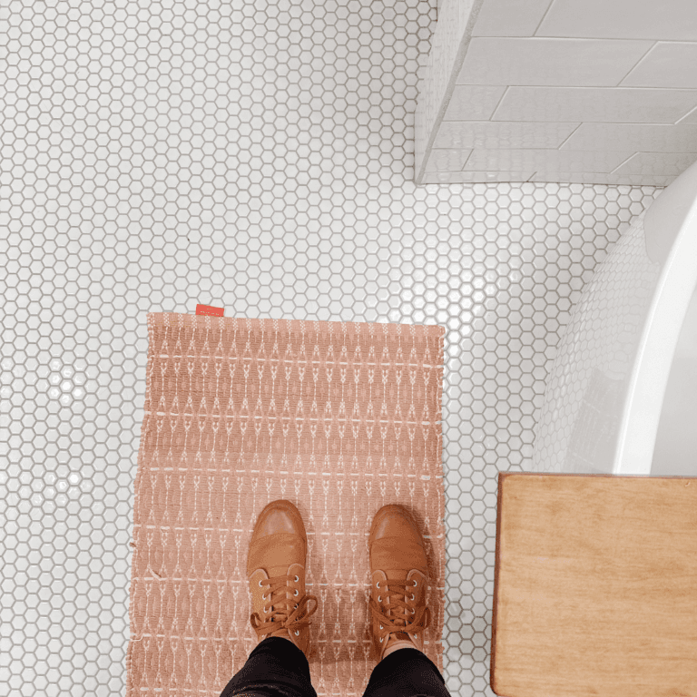 Pair of shoes standing in an all-white bathroom with white hex floor tile with an orange bath mat