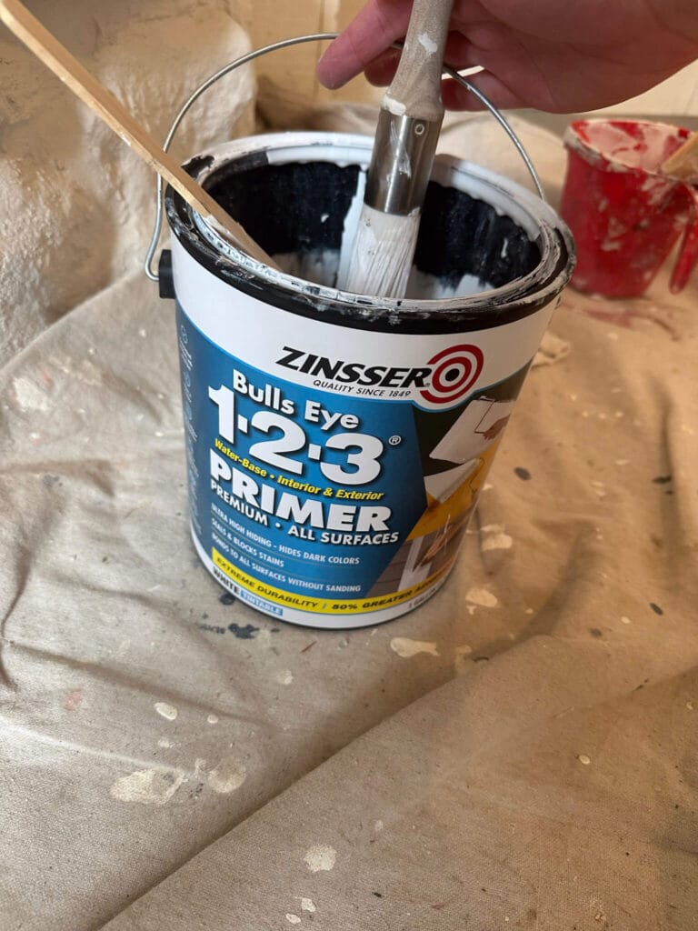 Open can of Zinsser 1-2-3 primer with a round brush being dipped into it next to a painted stone fireplace